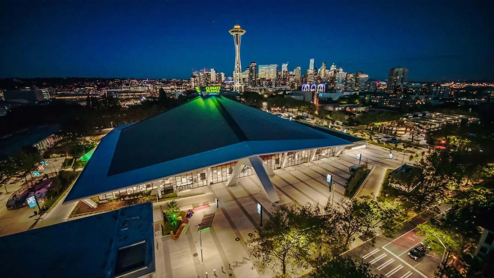 Aerial nighttime shot of Climate Pledge Arena in Seattle showing its distinctive, glowing, tent-like historic roof, with lit pedestrian plazas surrounding it and the illuminated Space Needle and Seattle skyline in the background.