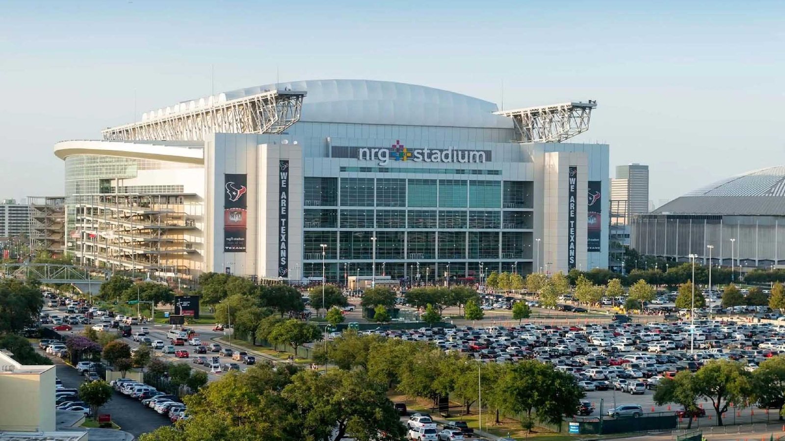 Daytime wide view of Reliant Stadium, pictured as NRG Stadium, highlighting its industrial glass facade and prominent exposed steel roof trusses, with the curved roof of the historic Houston Astrodome visible on the far right.